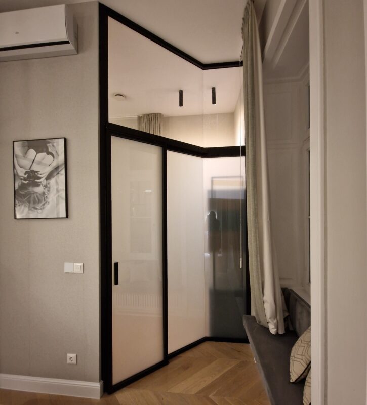 Corner of a modern bedroom with glass-front wardrobes, beige panels and dark trim, plus a cushioned bench with pillows by a window curtain.